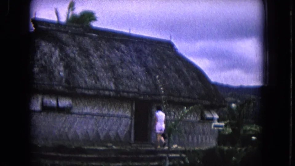 1968 Honolulu 8mm frame showing a person approaching the dark entrance of a large thatched hut with tropical greenery nearby.