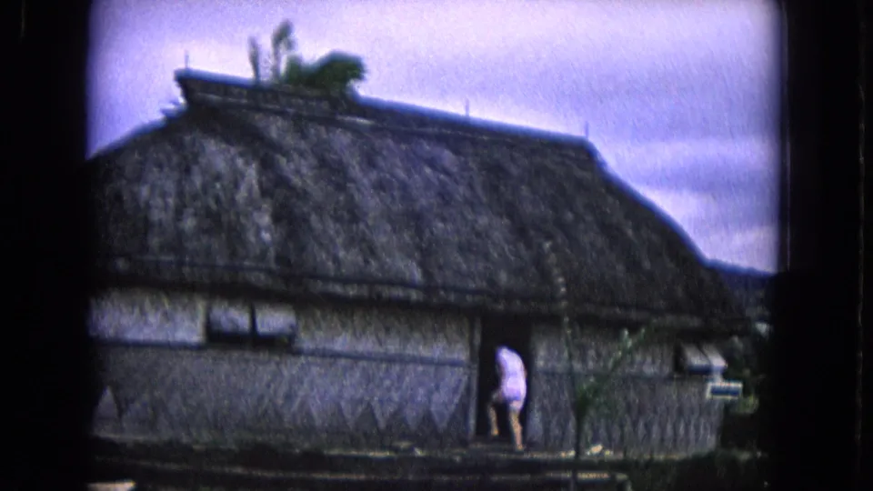 1968 Honolulu frame of a person entering a dark doorway in a thatched-roof hut, with tropical plants and cloudy sky visible.