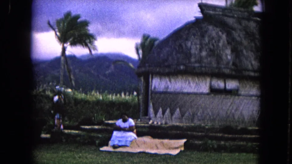 A woman in a light dress sits on a mat outside the thatched hut, with palm trees, grass, and low mountains visible in the background.
