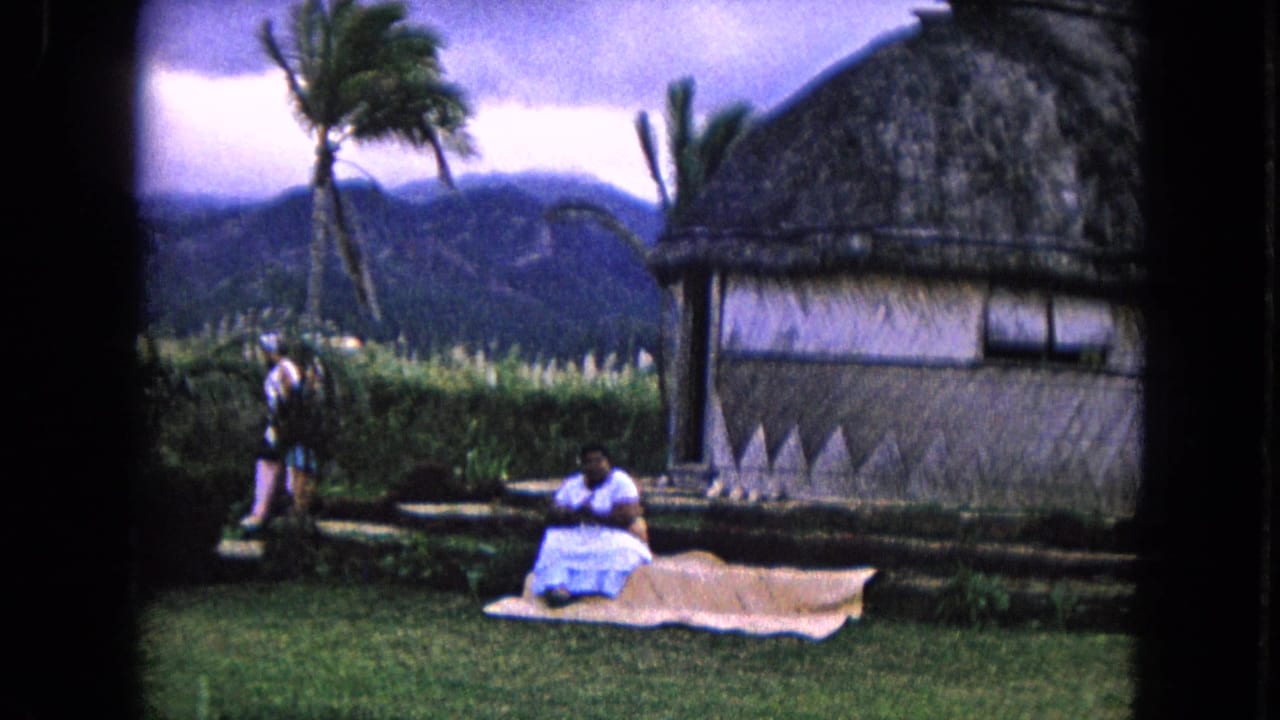 Woman in a white dress sits on a mat near a thatched hut with palms and mountains behind her.