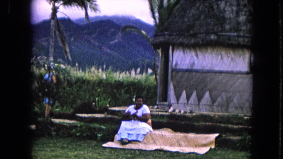 1968 Honolulu frame with the camera panned closer to a seated woman in a white dress beside greenery and a thatched building.