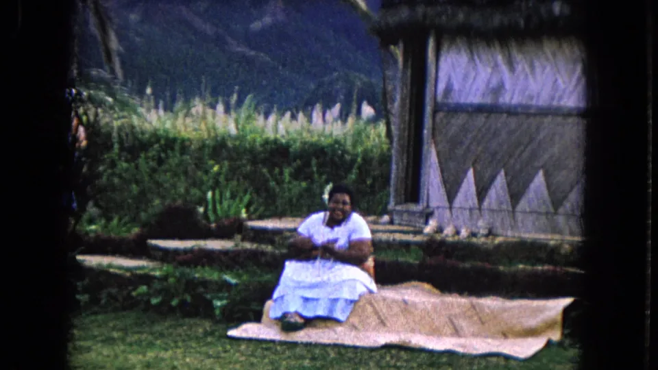 Closer 1968 Honolulu home-movie view of a smiling woman on a mat near garden plants, a thatched structure, and mountains.