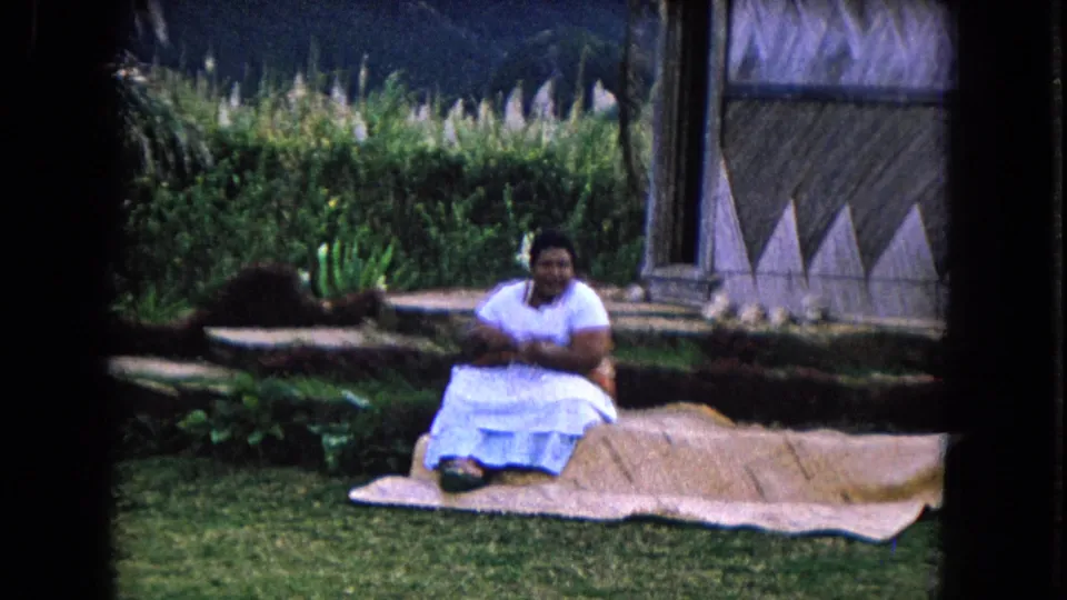 Final 1968 Honolulu frame shows the woman seated on a woven mat beside a thatched hut wall with dense greenery and mountains behind.