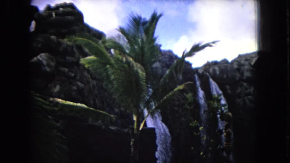 Soft 1968 Honolulu 8mm view of palm leaves, dark rock formations, and a waterfall against a bright sky.