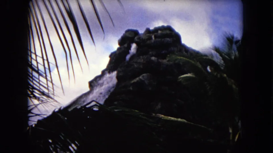 1968 Honolulu home-movie frame showing a dark artificial rock waterfall partly framed by palm fronds.