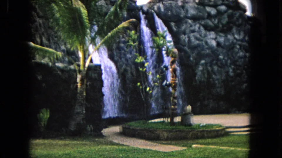 1968 Honolulu garden scene showing a waterfall, circular planter, lawn, walkway, palms, and a tiki-style carved figure.
