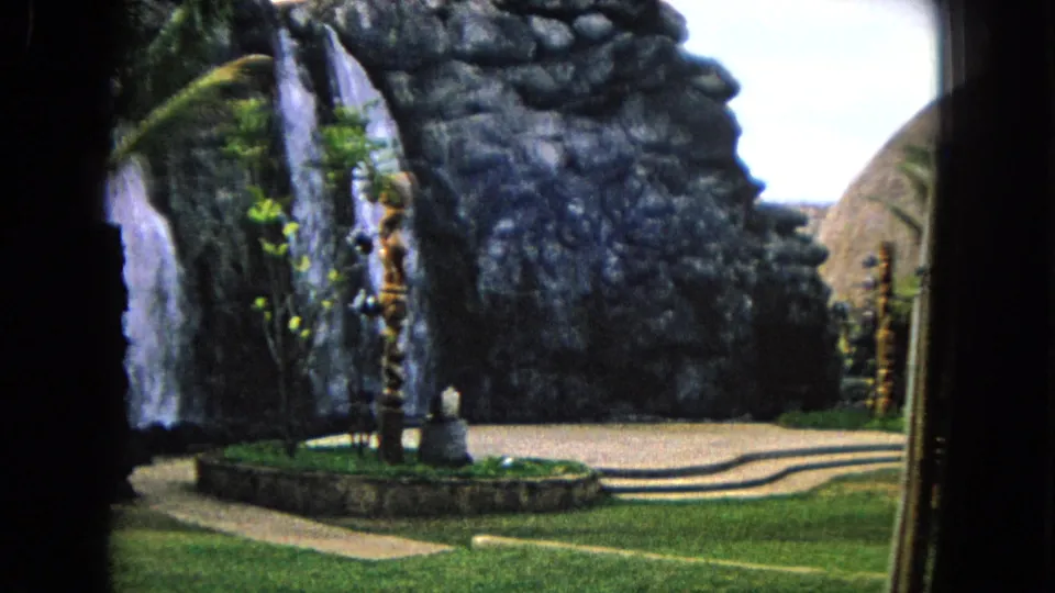 1968 Honolulu home-movie frame with a landscaped rock waterfall beside a lawn, walkway, palms, and circular planting bed.