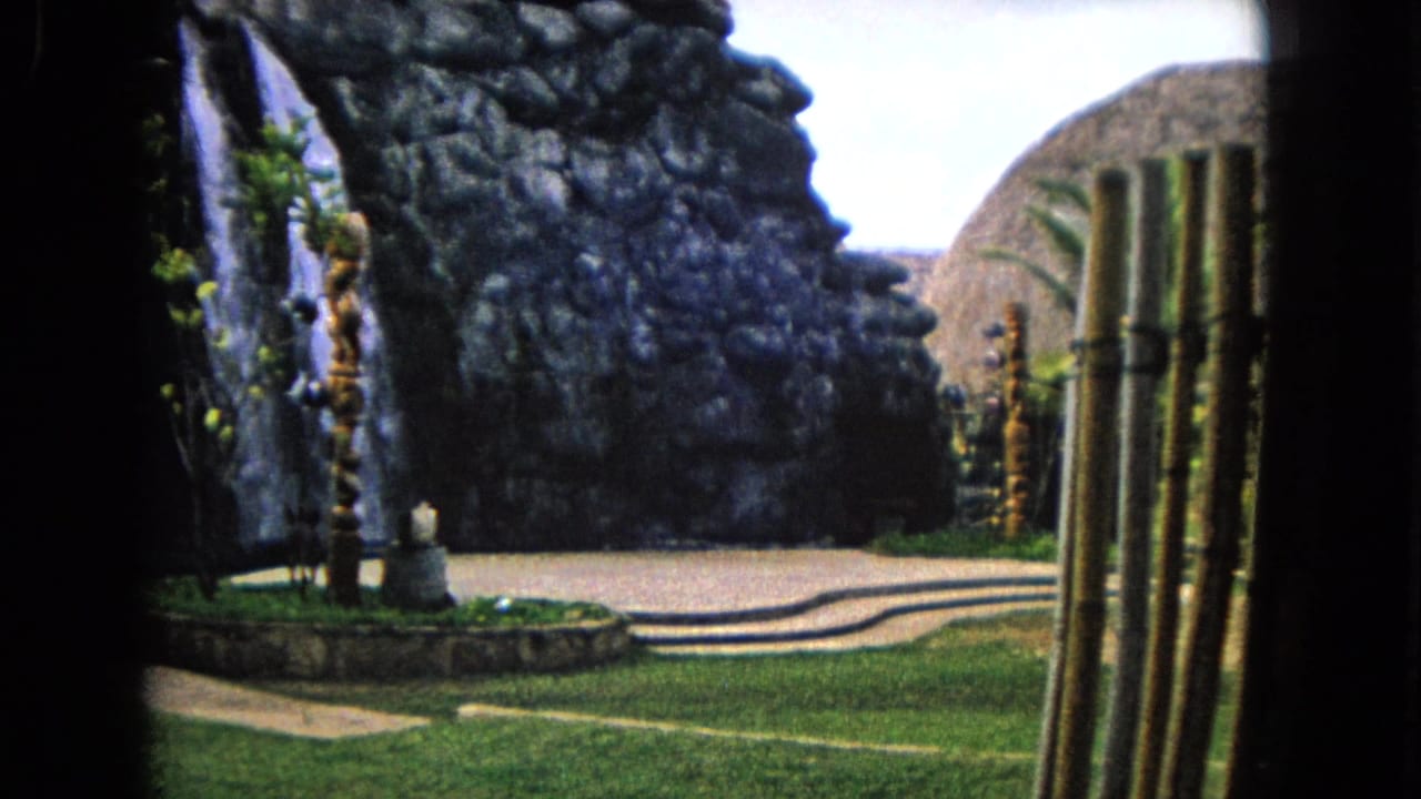 1968 Honolulu 8mm frame showing a landscaped tropical garden with a rock waterfall feature, walkway, grass, palms, and tiki-style wooden carvings.