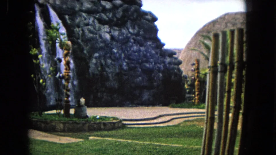 1968 Honolulu 8mm frame showing a landscaped tropical garden with a rock waterfall feature, walkway, grass, palms, and tiki-style wooden carvings.