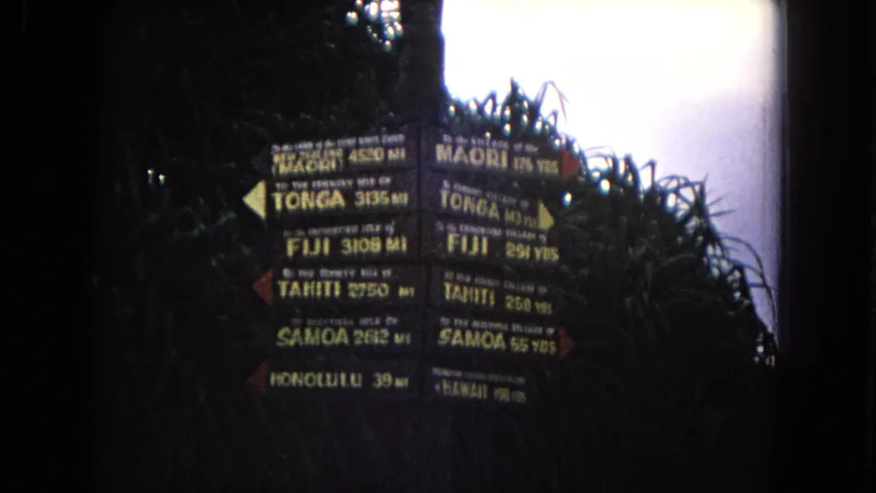 1968 Honolulu 8mm frame showing a weathered direction sign with Pacific island names and distance markings, filmed in shadow against foliage.