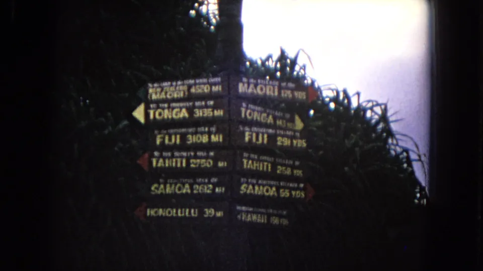 1968 Honolulu 8mm frame holding on the Pacific island distance signpost, with wooden arrows, plant shadows, and a pale overexposed sky edge.