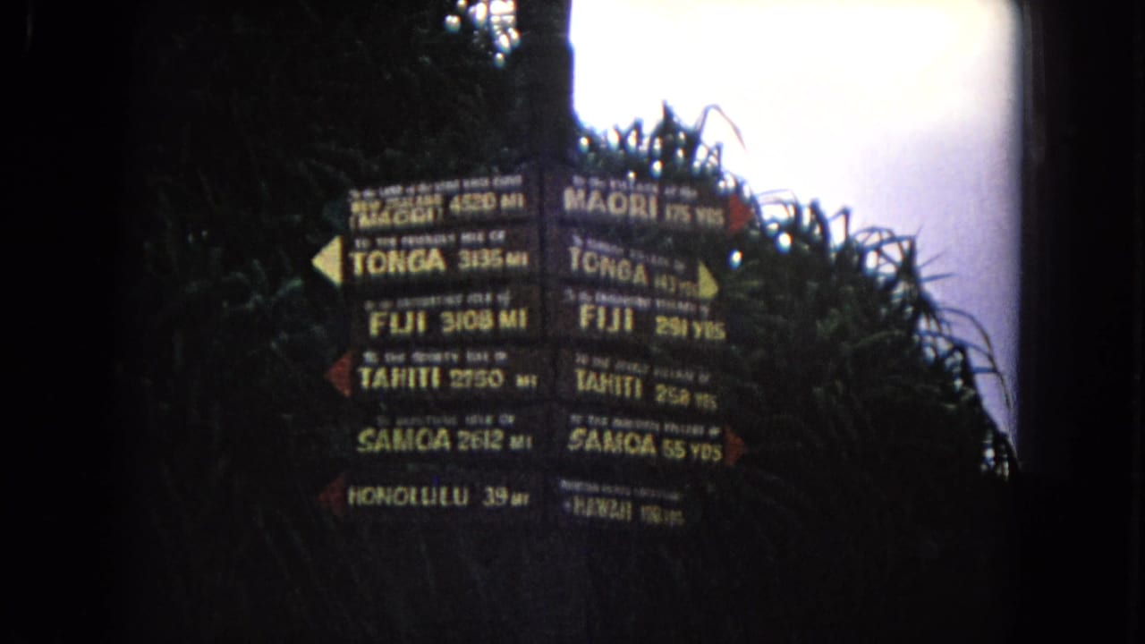 1968 Honolulu home-movie frame of a Pacific island directional signpost surrounded by dense foliage and bright sky.