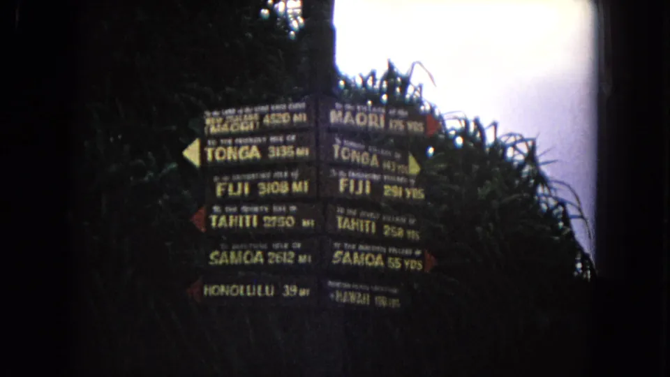 1968 Honolulu home-movie frame of a Pacific island directional signpost surrounded by dense foliage and bright sky.