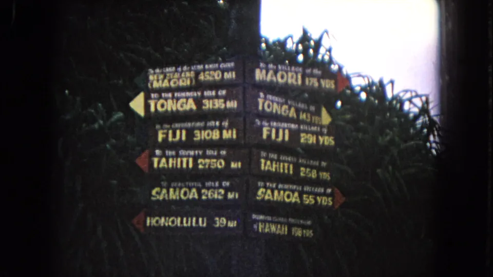 1968 Honolulu home-movie frame of the Pacific island signpost, with readable destination names and leafy shade.