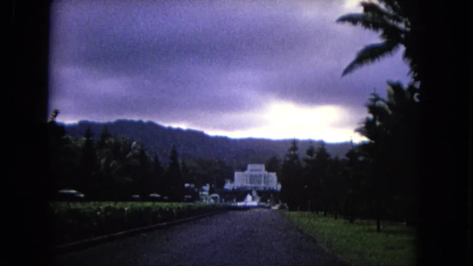 1968 Honolulu home-movie frame of a long landscaped approach toward a white building with mountains and heavy clouds behind it.