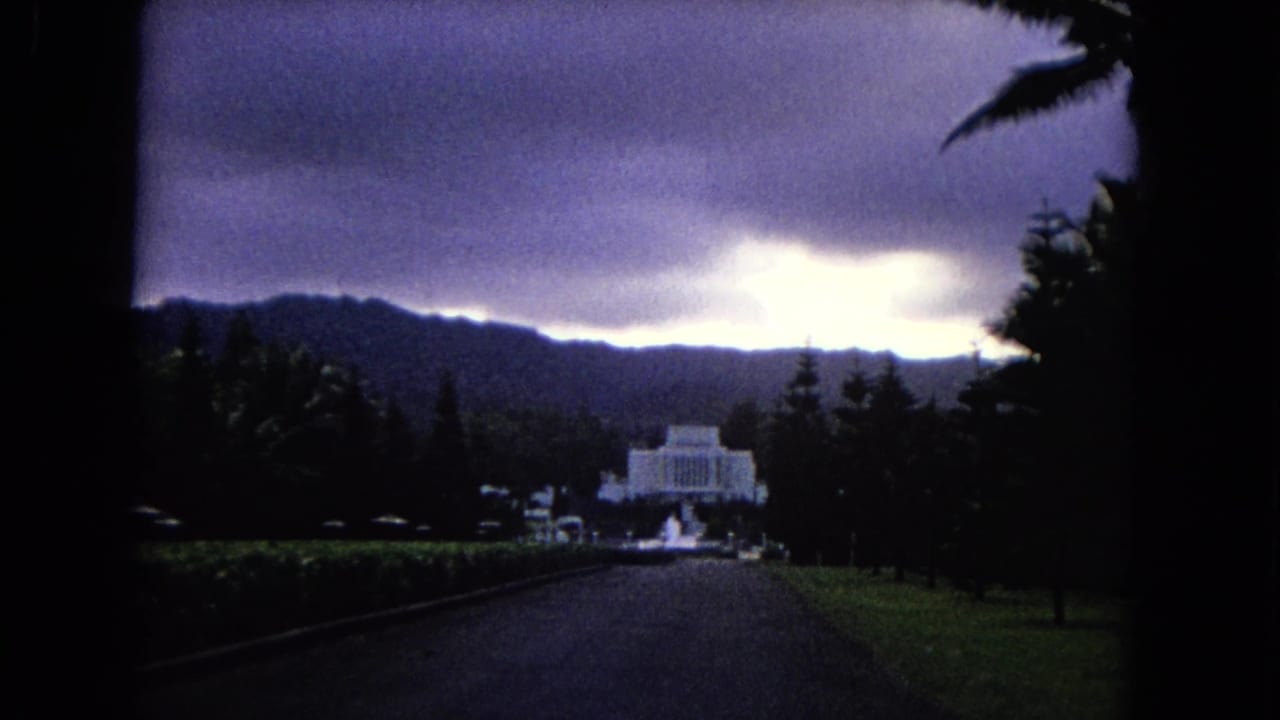 Long driveway leading toward a white temple-like building under dark hills and a bright stormy sky.