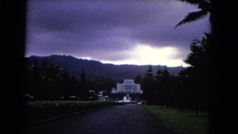 1968 Hawaii home-movie frame looking down a broad drive toward a distant white temple-like building framed by trees, hills, and storm clouds.