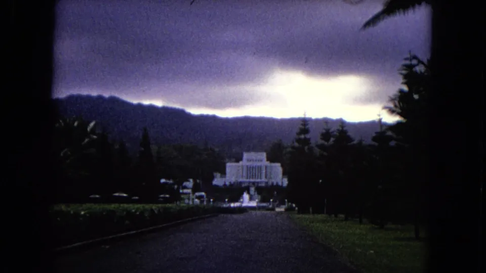 A 1968 Hawaii frame shows a white temple-like landmark beyond a dark driveway, with trees, small vehicles, hills, and a bright break in the clouds.