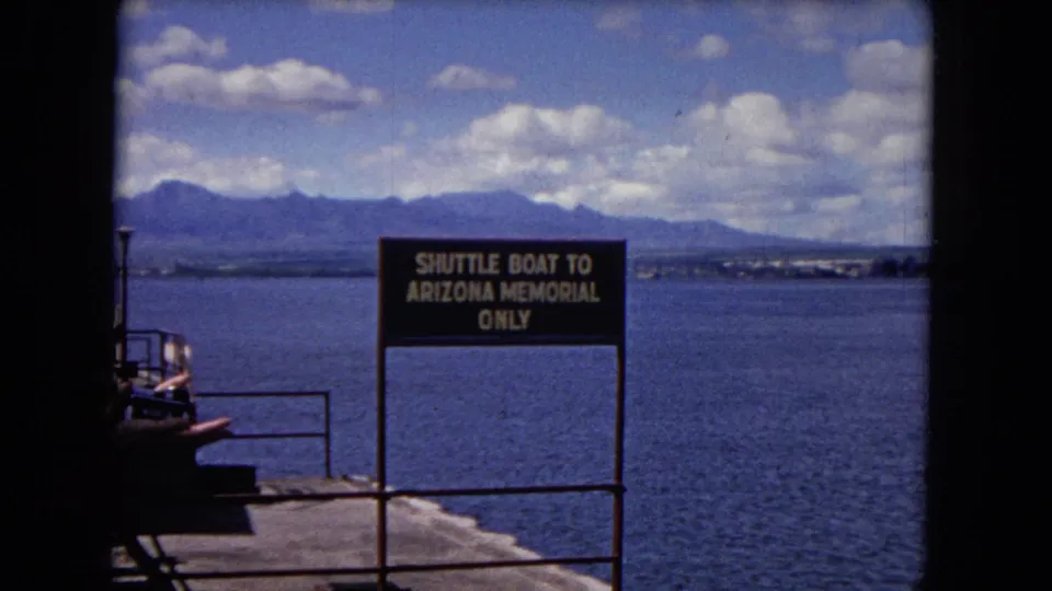 1968 Honolulu home-movie view of the Arizona Memorial shuttle sign near the pier, with harbor water, mountains, and a partial visitor visible at left.