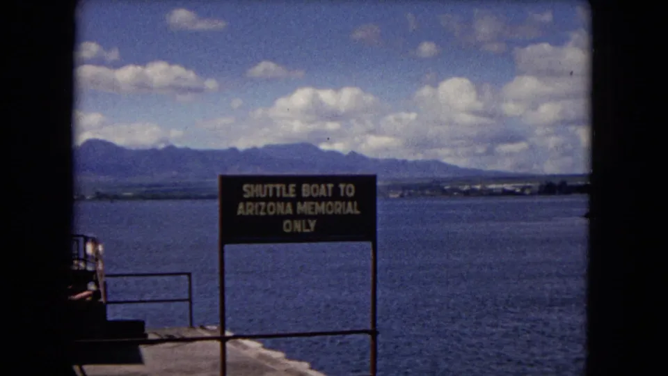 1968 Honolulu visitor-shot frame with the Arizona Memorial shuttle boat sign, pier railing, harbor water, and clouded mountain backdrop.