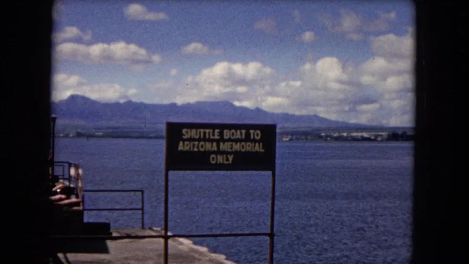 1968 Honolulu home-movie frame showing a pier-side Arizona Memorial shuttle sign against harbor water and the mountain landscape.