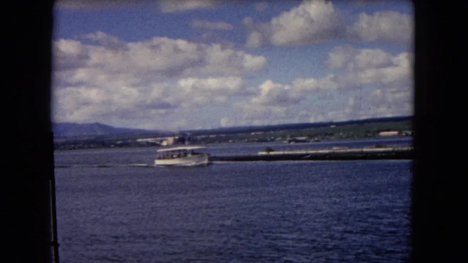 1968 Honolulu harbor view with a small white shuttle boat moving across blue water under a partly cloudy sky.