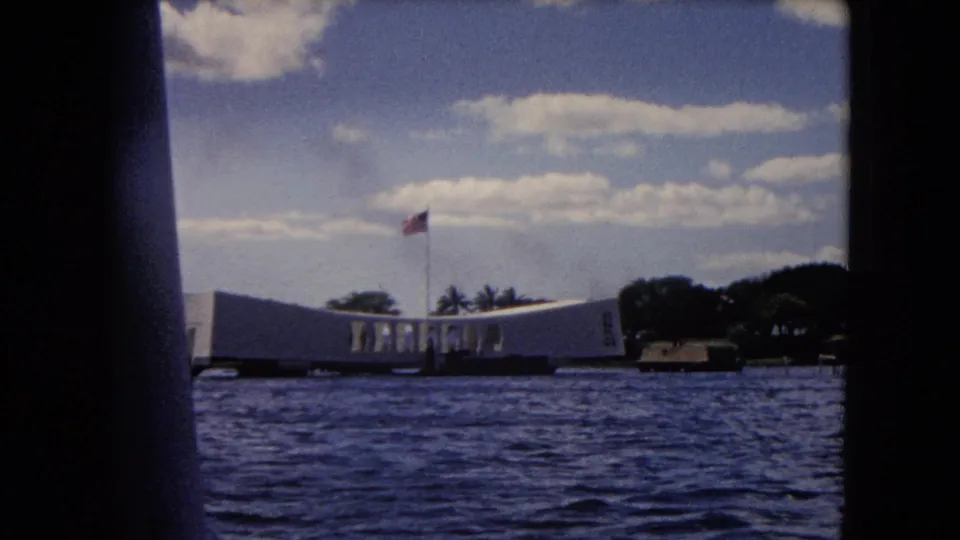 1968 water-level view of the USS Arizona Memorial, partly framed by a dark boat edge, with sky and trees in the background.