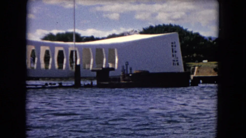 Close 8mm view of the USS Arizona Memorial from the water, with the curved wall, openings, and trees behind it.