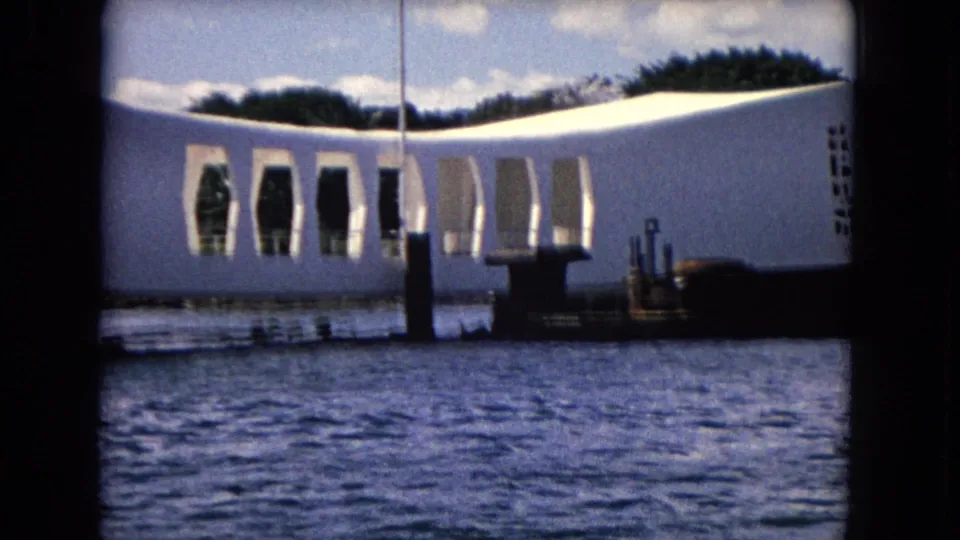 Pearl Harbor view of the USS Arizona Memorial from the water, showing its white curved form, open arches, dark posts, and trees beyond.