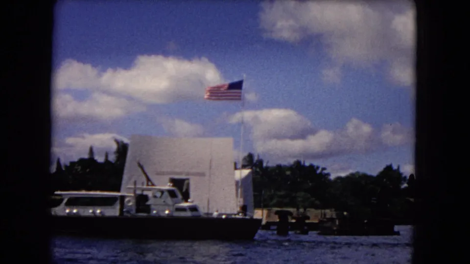 1968 visitor-shot Pearl Harbor frame with an American flag, tour boat, white memorial structure, dock, water, and cloudy sky.