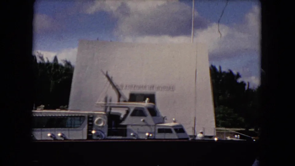 USS Arizona Memorial at Pearl Harbor with a docked boat in front, faint memorial lettering, water, trees, and vertical film wear visible.