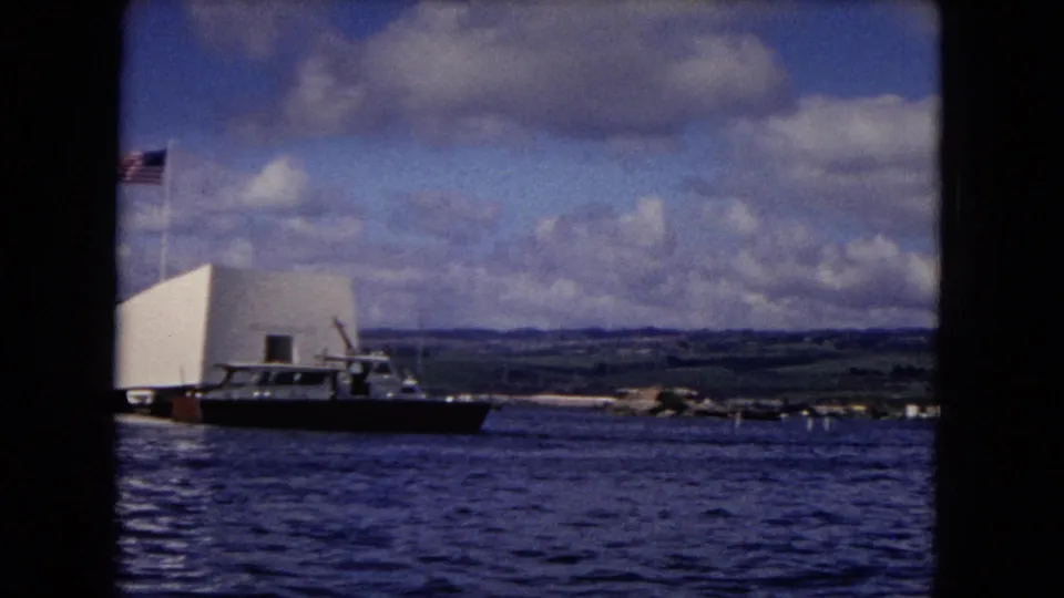 1968 Pearl Harbor frame showing the USS Arizona Memorial beside harbor water, with flag, boat, clouds, and distant landscape.