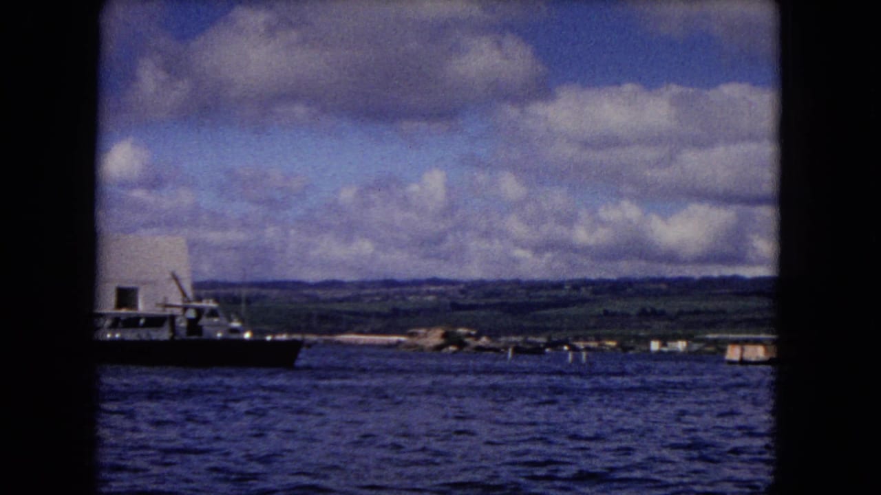 Naval ship on blue water near a low Honolulu shoreline under cloudy sky.