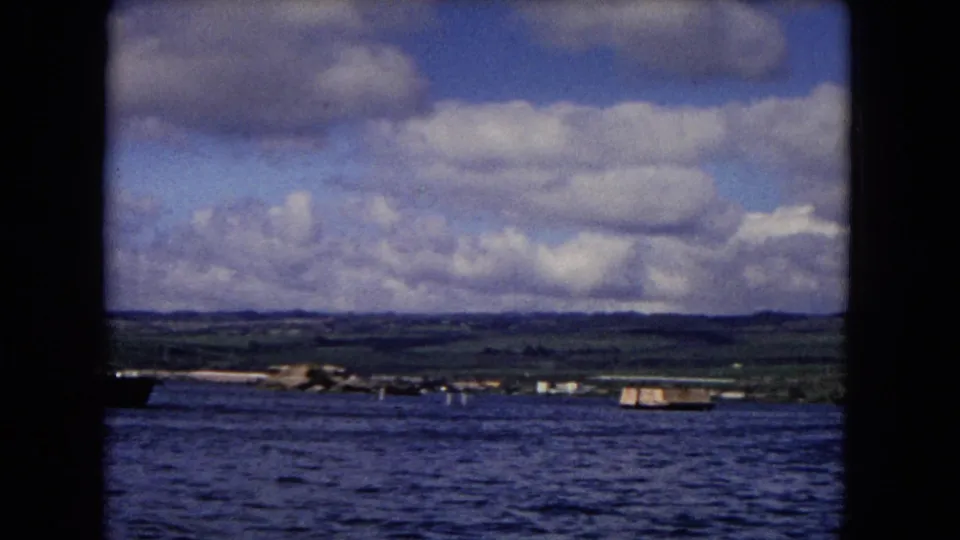 Honolulu coastal view from a moving vessel, showing water, low shore, small craft, hills, and cloudy sky.