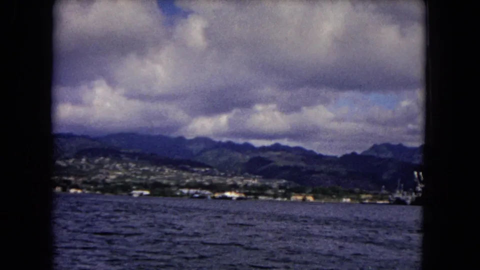 Honolulu shoreline seen across open water, with mountains, scattered buildings, clouds, and a cropped vessel at right.