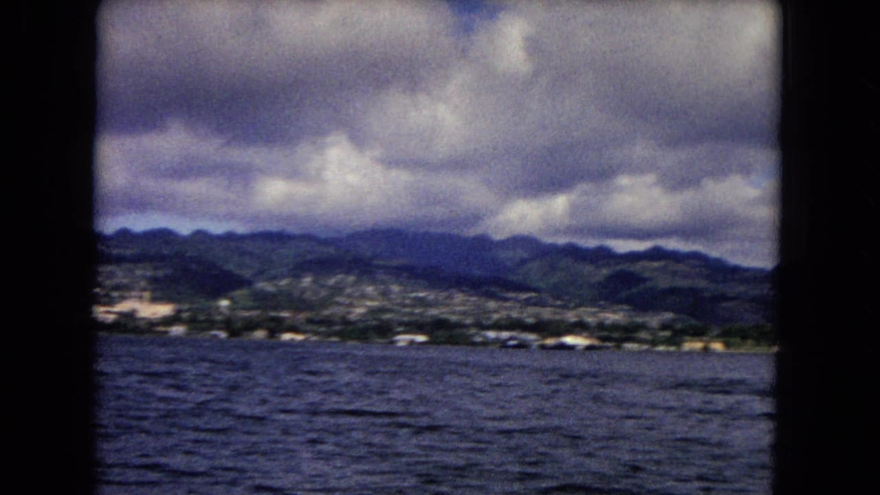 1968 8mm view of Honolulu shoreline, green mountains, cloudy sky, and choppy water framed by a boat window.