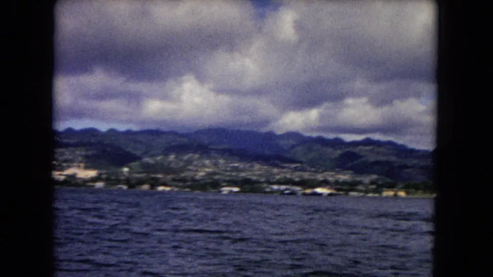 1968 8mm view of Honolulu shoreline, green mountains, cloudy sky, and choppy water framed by a boat window.