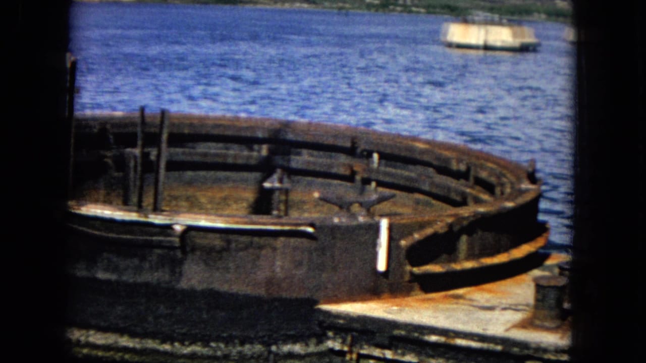 Rusted circular metal foundation beside blue harbor water with distant shoreline.