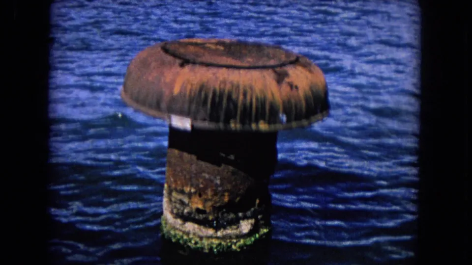 1968 Honolulu harbor footage shows a rusted cylindrical buoy or piling with barnacle staining rising from blue seawater.