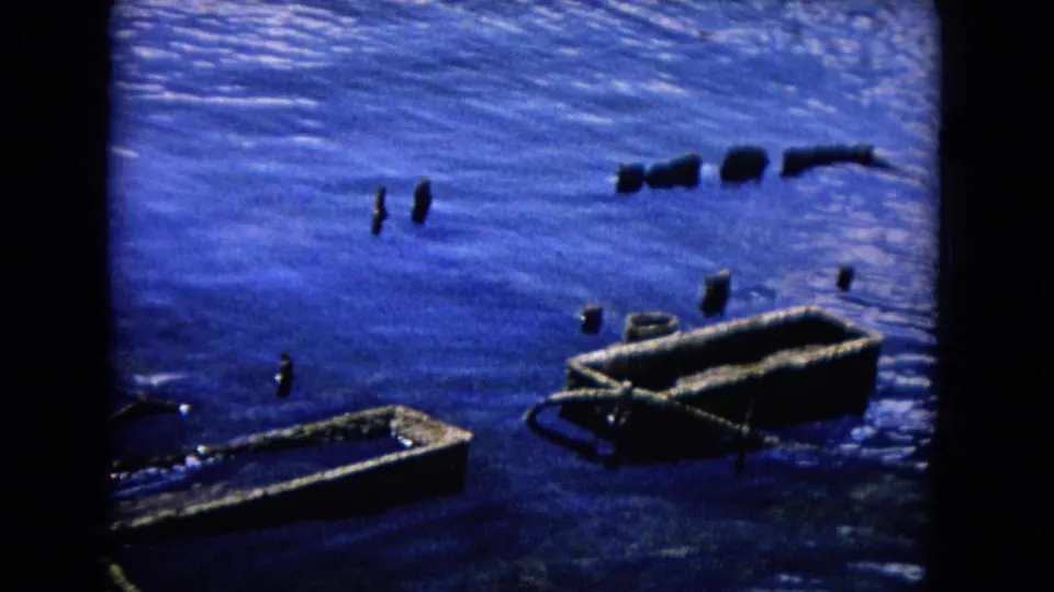 1968 Honolulu waterfront frame showing empty hulls, scattered posts, and fragments of a damaged dock in blue water.