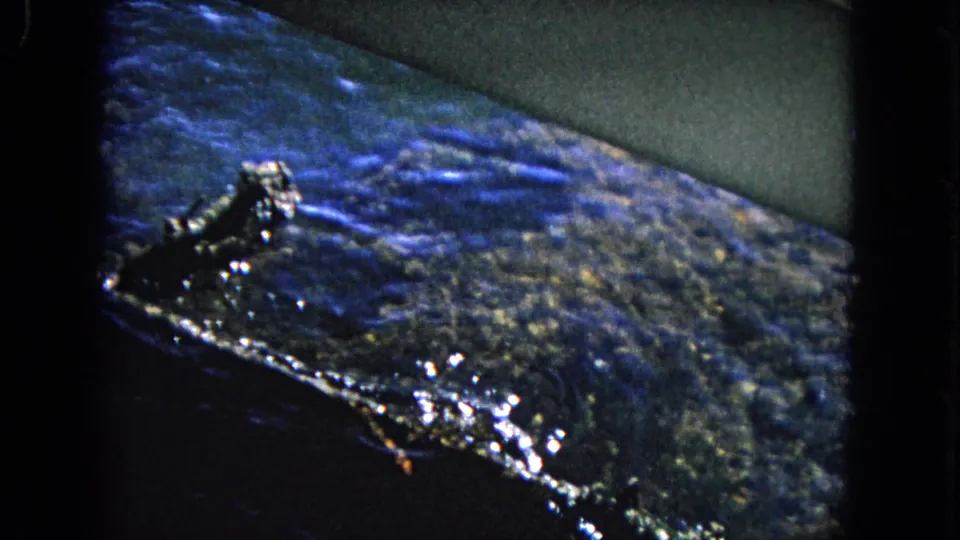 Grainy 1968 Honolulu home-movie frame showing blue water, a dark shoreline or harbor edge, and scattered bright reflections.