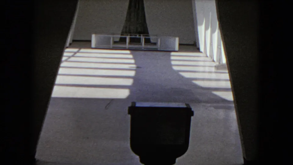 1968 Honolulu home-movie frame of a bright modernist corridor with a dark object near the camera and a tall central structure beyond.