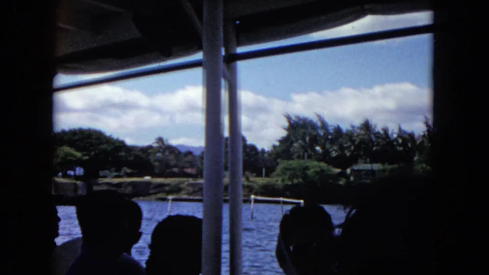 Passengers in silhouette look out from a Honolulu boat toward calm water, tropical trees, distant hills, and clouds.