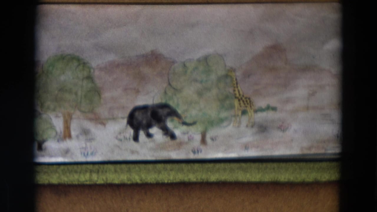 1968 Lahaina Maui home-movie frame showing a handmade painted savanna backdrop with an elephant, giraffe, trees, and hills.