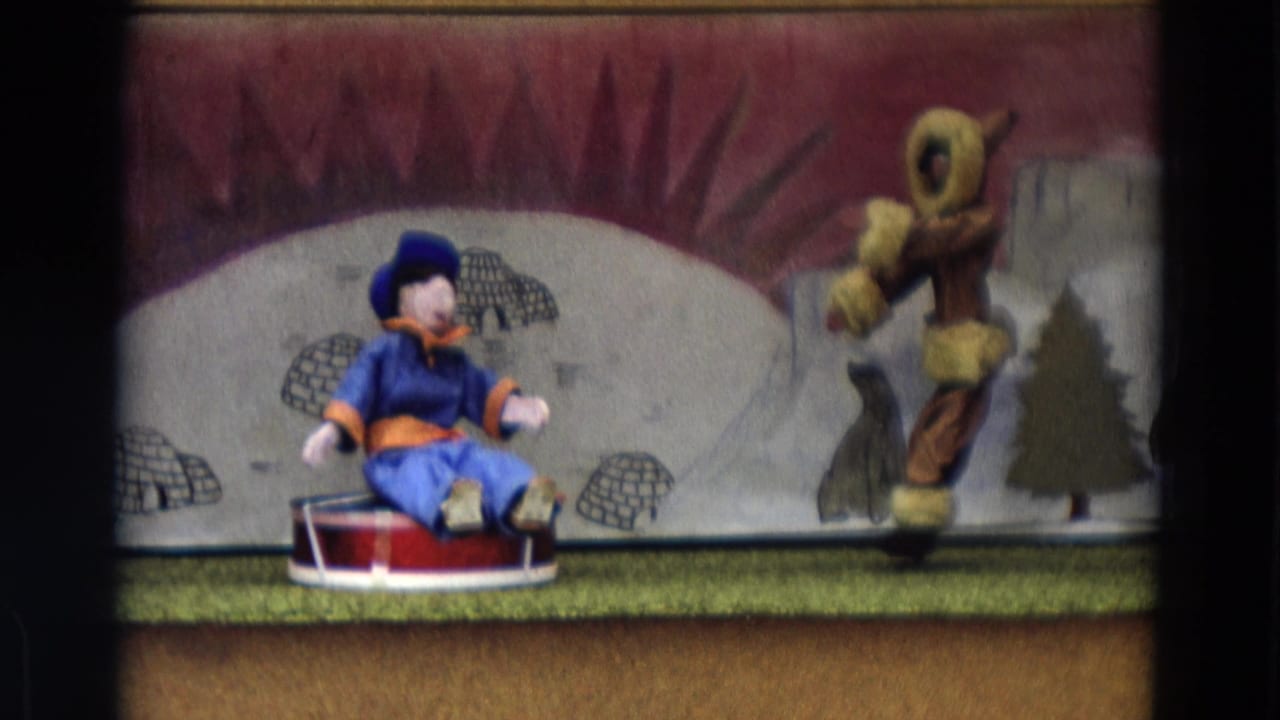 Blue puppet seated on a red sled beside a larger fur-trimmed puppet on an arctic stage backdrop.