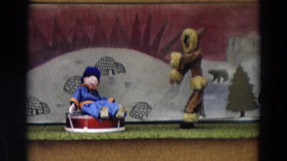 Home-movie frame from 1968 Lahaina Maui showing a blue-costumed puppet on a sled and a larger puppet on an arctic-themed stage.