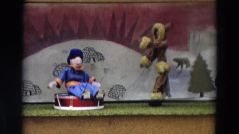 1968 Lahaina Maui 8mm frame of a staged string puppet scene with a blue sled puppet and a taller fur-trimmed figure before arctic scenery.