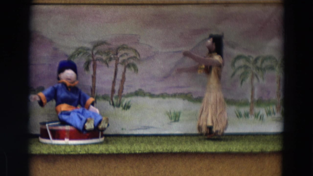 1968 Lahaina Maui 8mm frame with a seated puppet, dancer figure, and painted palm backdrop on a small stage.