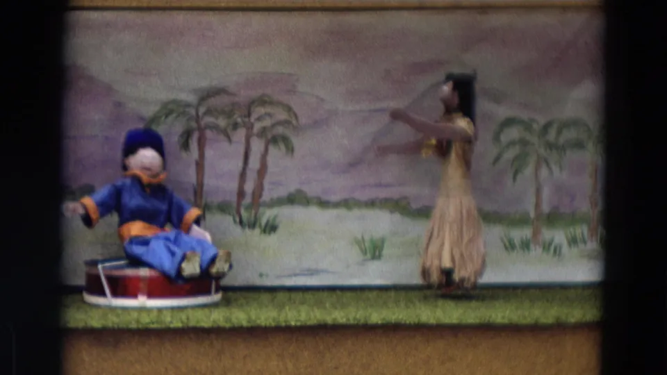 1968 Lahaina Maui 8mm frame with a seated puppet, dancer figure, and painted palm backdrop on a small stage.