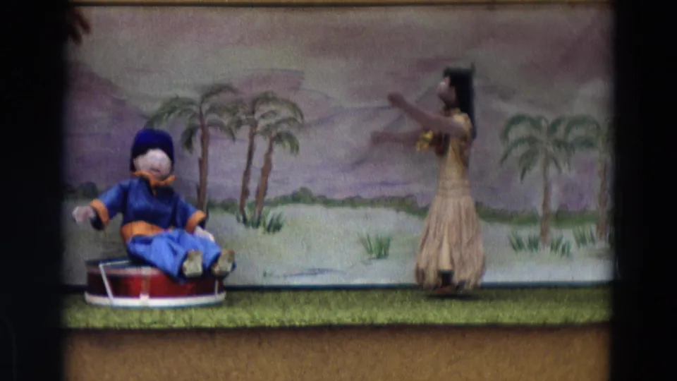 1968 8mm Lahaina Maui frame showing a small puppet-stage tableau with seated puppet, dancer figure, and palm backdrop.